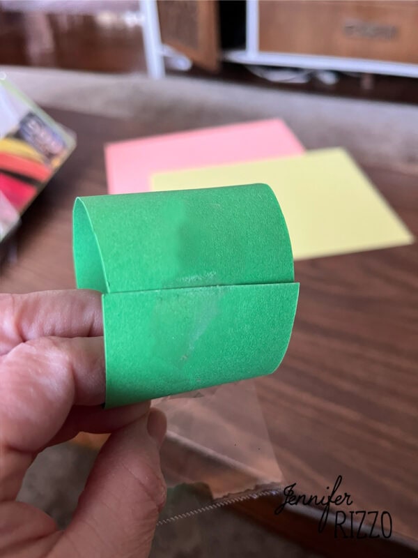 how to make an easy construction paper chain 4 a hand holds a loop made from green construction paper; colored paper sheets are on a wooden table in the background.