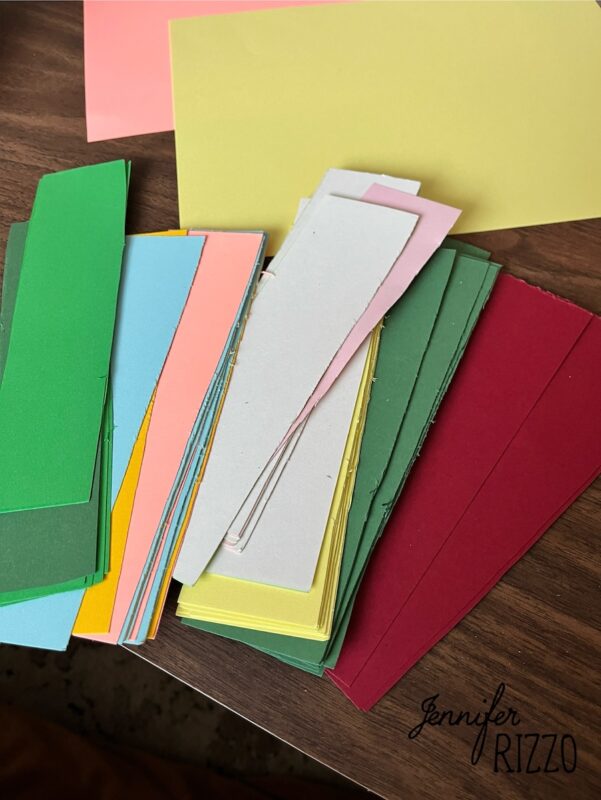 how to make an easy construction paper chain 3 a stack of colorful paper strips and sheets, including green, yellow, blue, pink, gray, and red, arranged on a wooden surface. the name "jennifer rizzo" is visible in one corner.