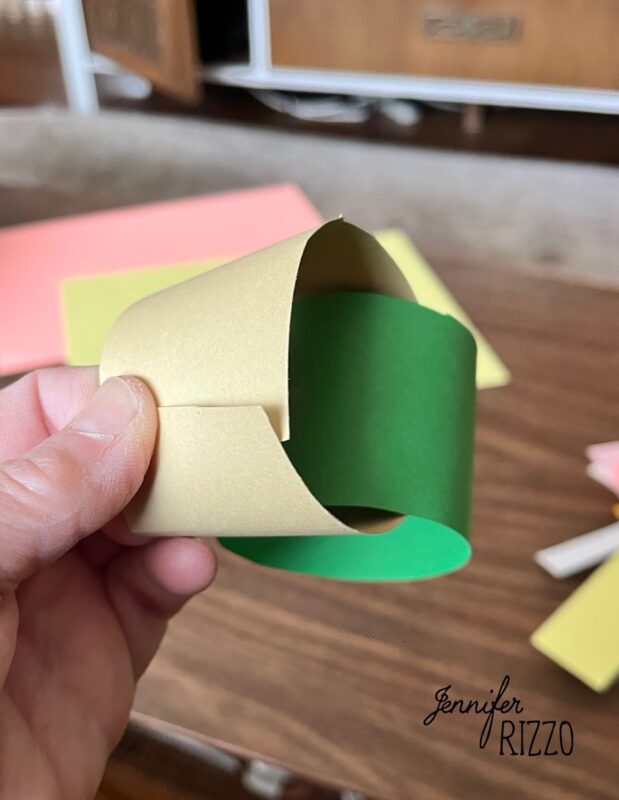 how to make an easy construction paper chain 5 a hand holds two interlocking paper loops, one gold and one green, on a wooden table with sheets of colored paper in the background.
