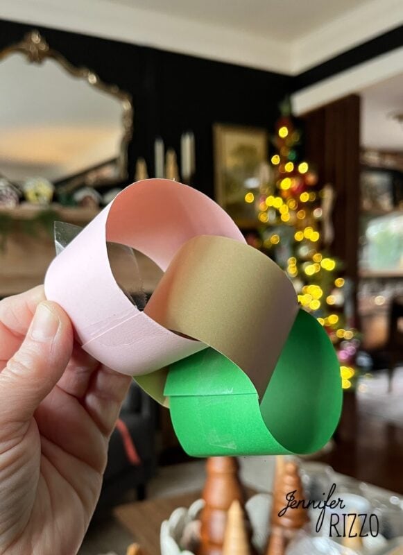 how to make an easy construction paper chain 6 a hand holds a three-link paper chain in pink, gold, and green with a christmas tree and holiday decor in the background.