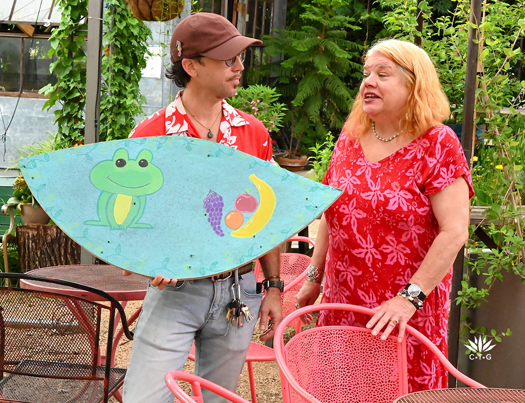 prairie up benjamin vogt new episode 10 young man holding brightly painted wood of a frog and fruit; showing it to bonnie cullum