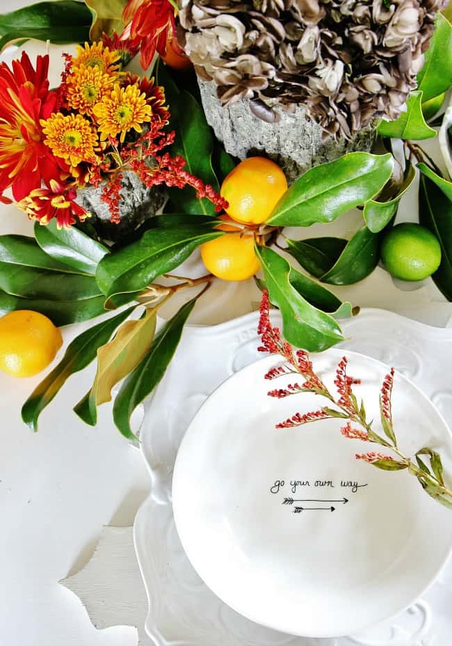 five fun family games to play this thanksgiving 17 a festive table setting featuring a white plate with the phrase "go your own way" inscribed on it, surrounded by vibrant flowers, citrus fruits, and a decorative rock vessel with mushrooms.