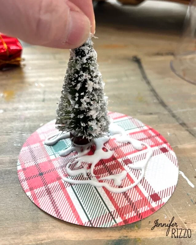 mini snow globe cloches from plastic wine glasses 4 a hand places a small, snow-covered artificial tree onto a plaid-patterned circle with visible white glue underneath.
