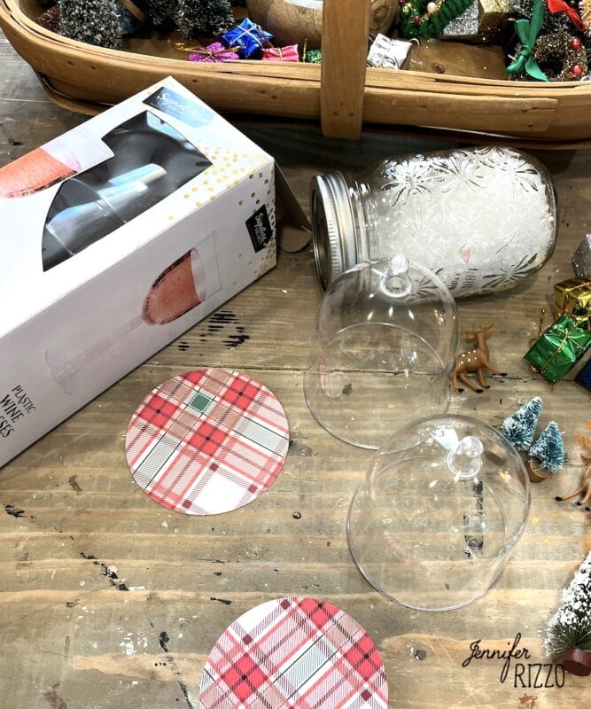 mini snow globe cloches from plastic wine glasses 1 two glass domes, plaid coasters, a box labeled "plastic wine glasses," a snow-filled jar, and mini holiday decorations on a wooden table. mini snow globe cloches from plastic wine glasses