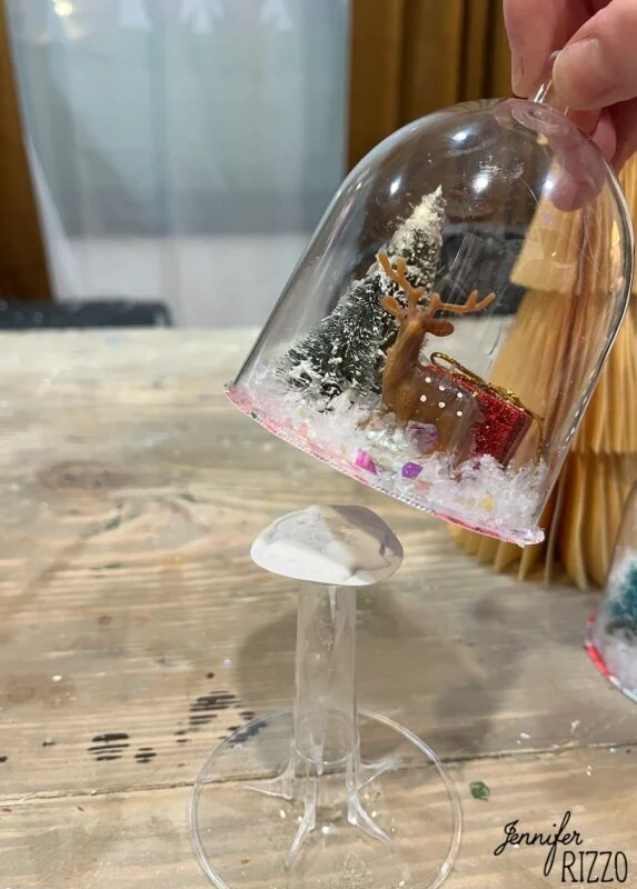 mini snow globe cloches from plastic wine glasses 8 a hand holds an upside-down wine glass with a miniature reindeer, tree, and gift scene inside, displayed on a wooden table.