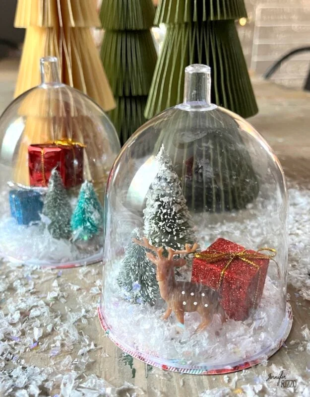 mini snow globe cloches from plastic wine glasses 10 miniature holiday scene with a plastic dome covering a small deer, trees, artificial snow, and red gift boxes; folded paper trees in the background to make mini snow globe cloches from plastic wine glasses
