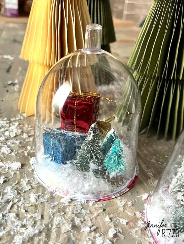 mini snow globe cloches from plastic wine glasses 12 a small holiday snow globe with miniature trees, gift boxes, artificial snow, and paper trees in the background.