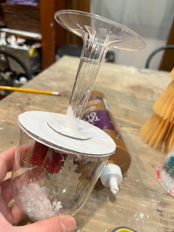 mini snow globe cloches from plastic wine glasses 9 a hand holds an upside-down wine glass with white cardboard glued to the base, containing small holiday decorations and faux snow inside the glass.
