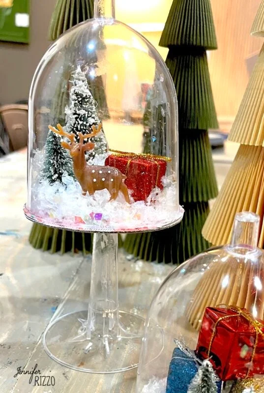 mini snow globe cloches from plastic wine glasses 11 a small deer figurine, gift boxes, and snowy trees are displayed inside an upside-down wine glass, creating a winter scene decoration.