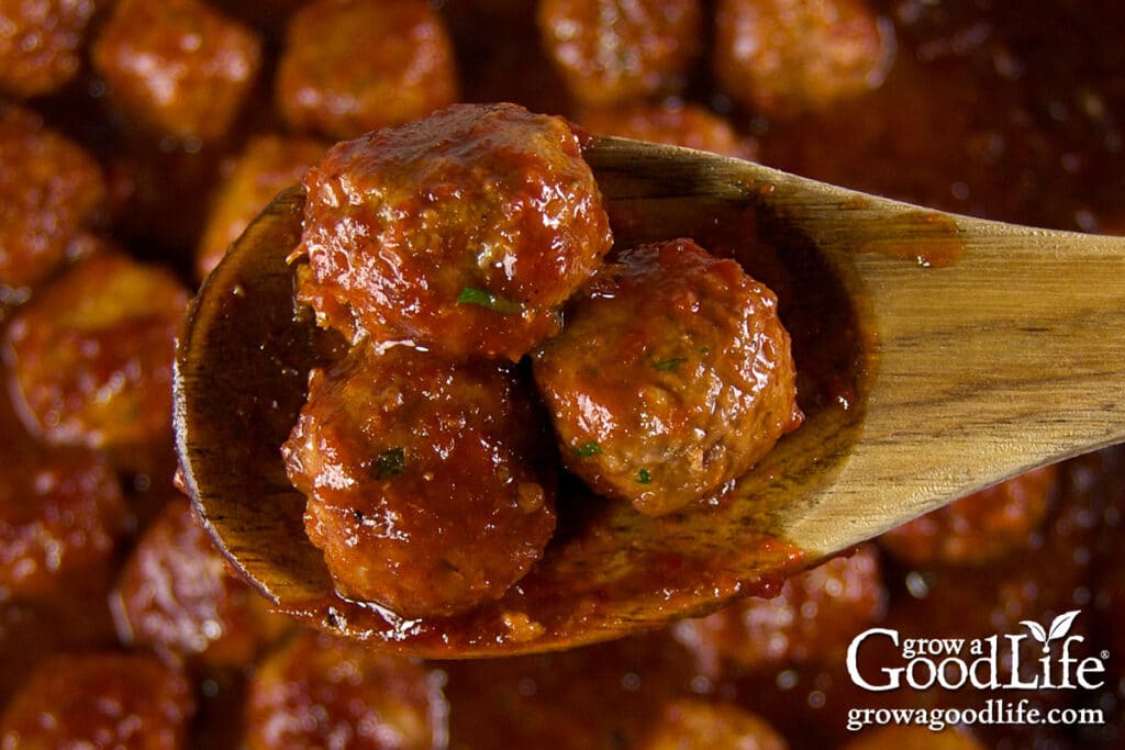 close-up of a wooden spoon lifting three saucy barbecue meatballs from the slow cooker.
