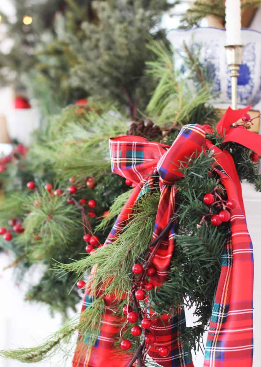 a festive holiday wreath made of pine branches and red berries, decorated with a red plaid ribbon, hangs against a white background with greenery and decorative items blurred in the background.