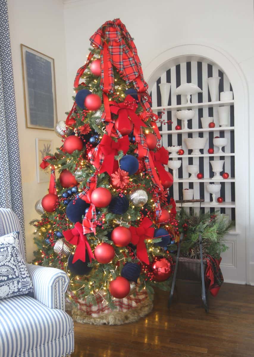 a decorated christmas tree with large red and navy blue ornaments, plaid ribbons, and red bows stands beside a white cabinet with festive decorations and greenery. warm white lights are strung on the tree.