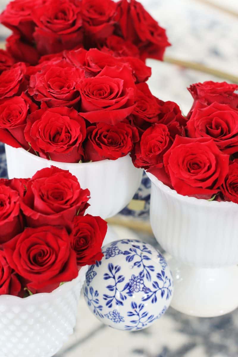 three white vases filled with vibrant red roses are arranged on a table. in front of them is a small decorative white sphere with a blue floral pattern.