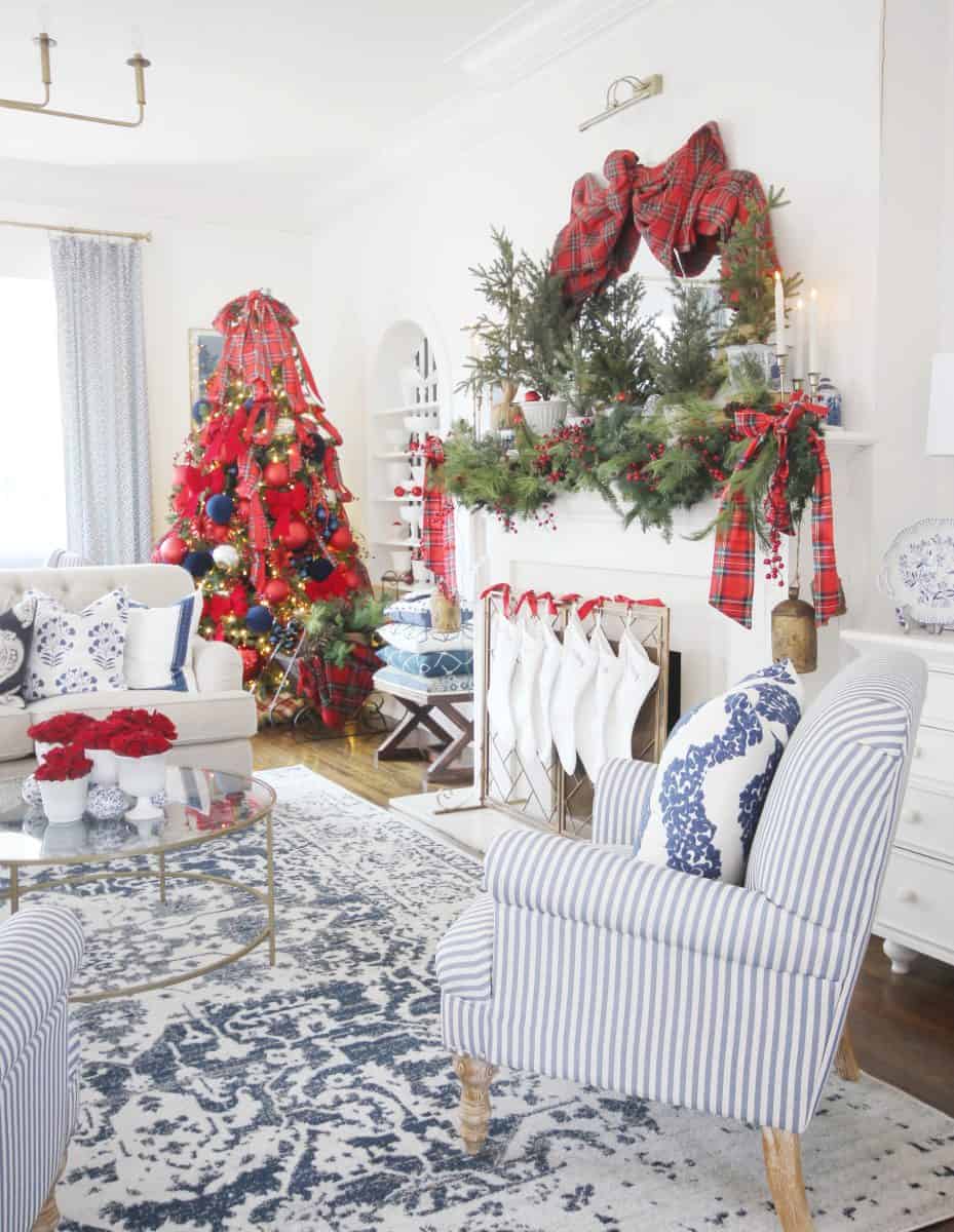 a bright living room decorated for christmas with a large red-themed tree, a mantel adorned with garland and plaid bows, stockings hanging, blue and white accents, and striped armchairs.