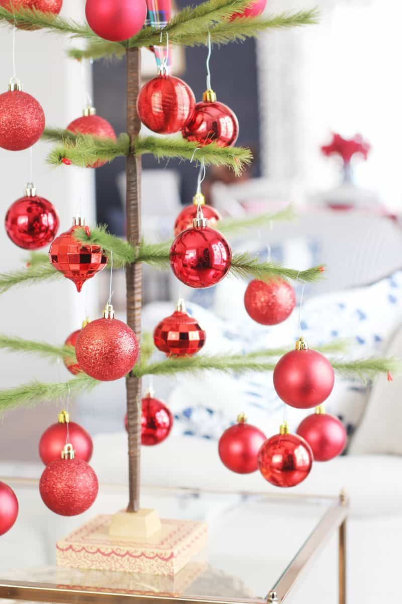 a minimalist christmas tree with sparse green branches decorated with shiny and glittery red ornaments stands on a glass table in a bright, modern living room.