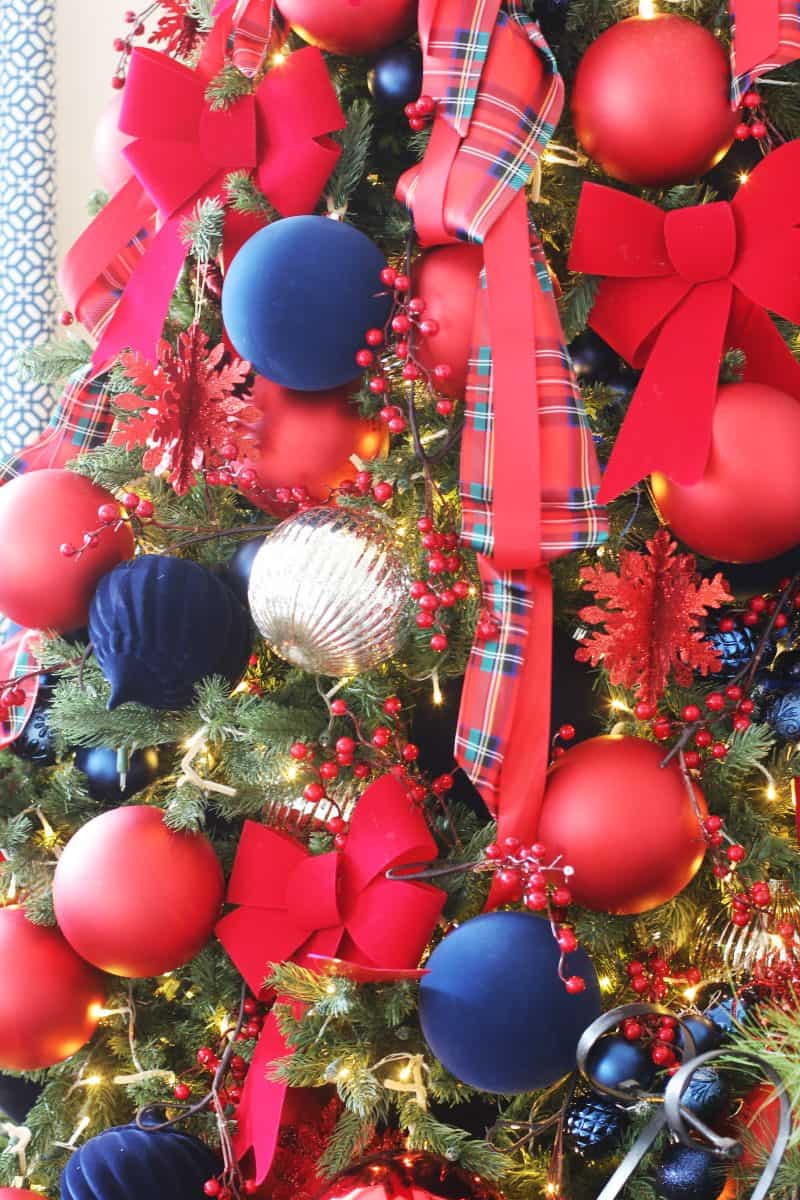 a christmas tree decorated with large red and navy blue ornaments, red bows, plaid ribbon, gold ornaments, glittery red snowflakes, and small string lights.