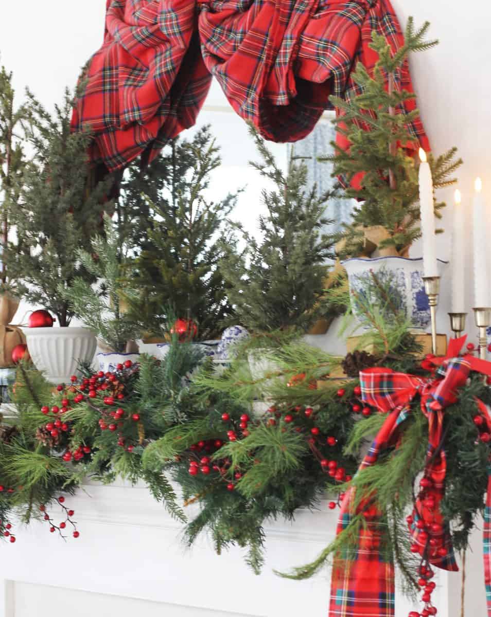 a festive mantel decorated with pine branches, red berries, plaid ribbons, small evergreen trees in blue and white vases, candles, and ornaments, with a red plaid fabric draped above.