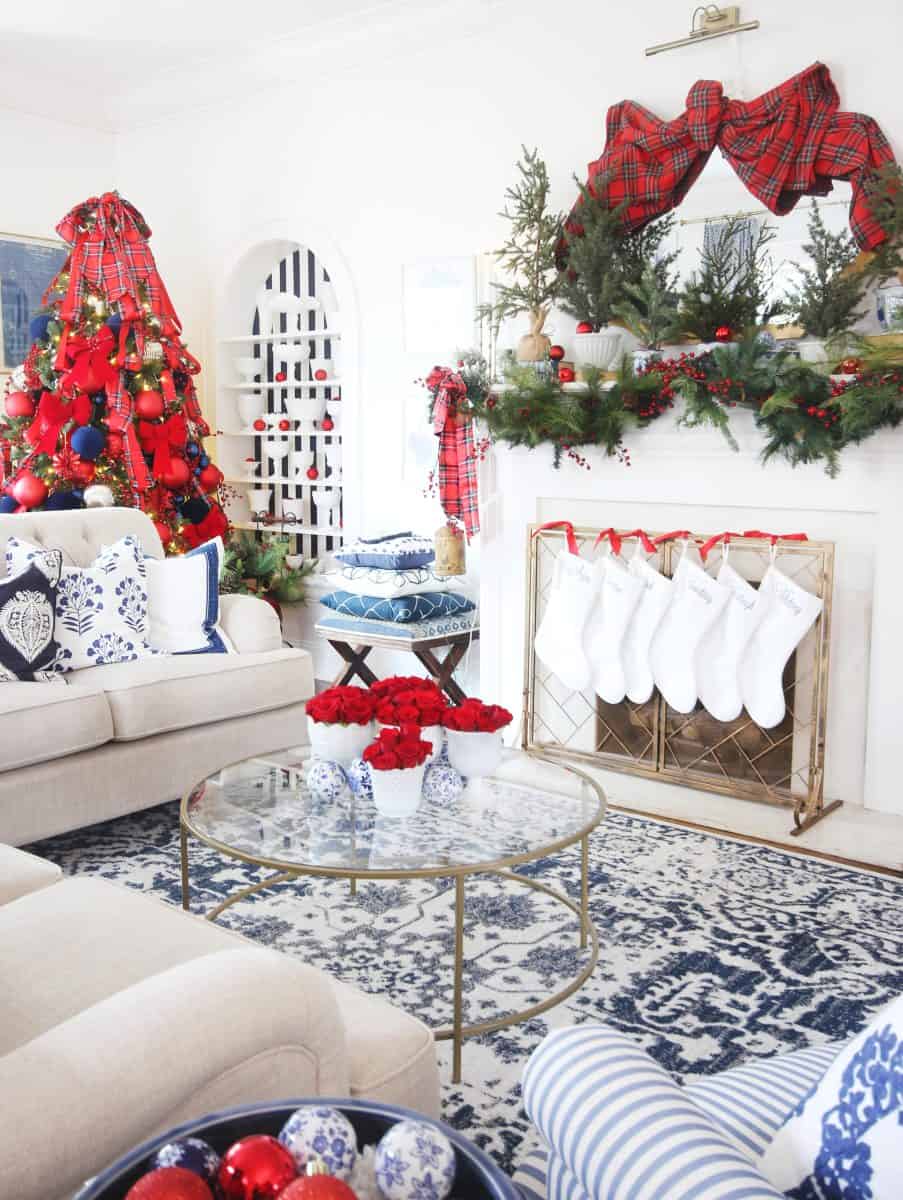 bright living room decorated for christmas with a red and blue theme, featuring a red christmas tree, stockings on a white fireplace, plaid ribbon wreath, and festive accents on a glass coffee table and furniture.