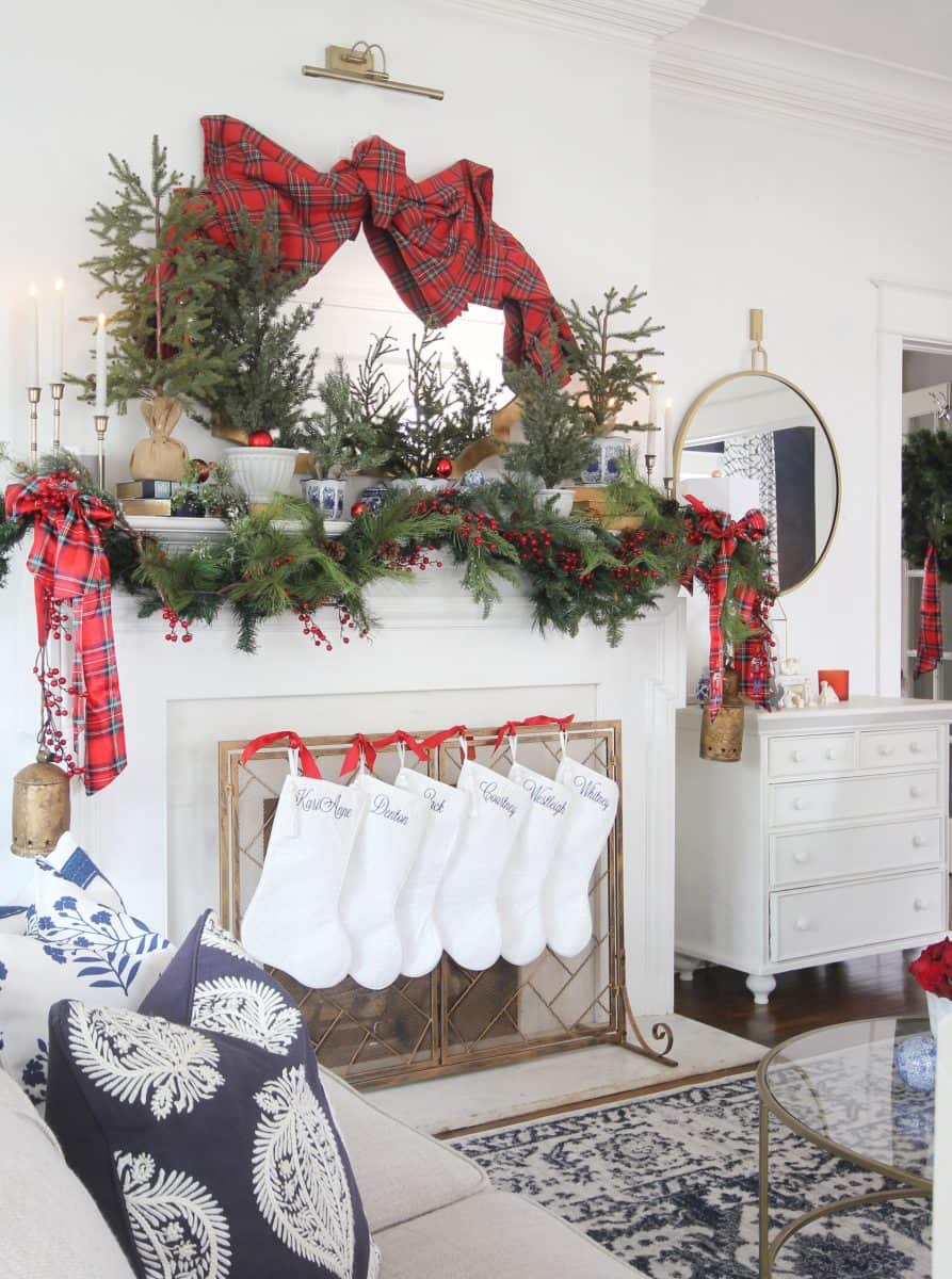 a festive living room with a white fireplace decorated with pine garlands, red berries, plaid bows, and mini christmas trees. five white stockings hang from the mantel, and a mirror and candles sit on top.