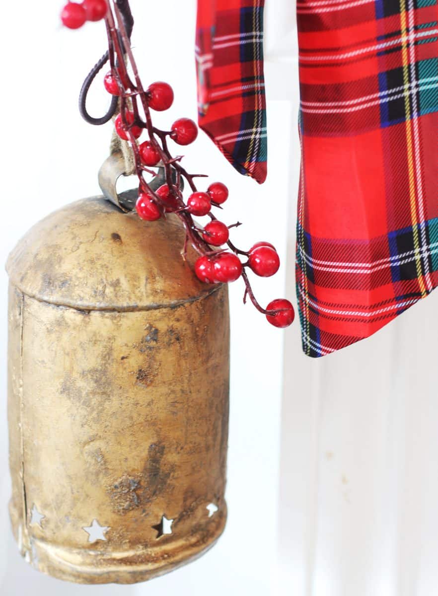 a large rustic gold bell decorated with red berries hangs next to a red plaid ribbon against a white background.