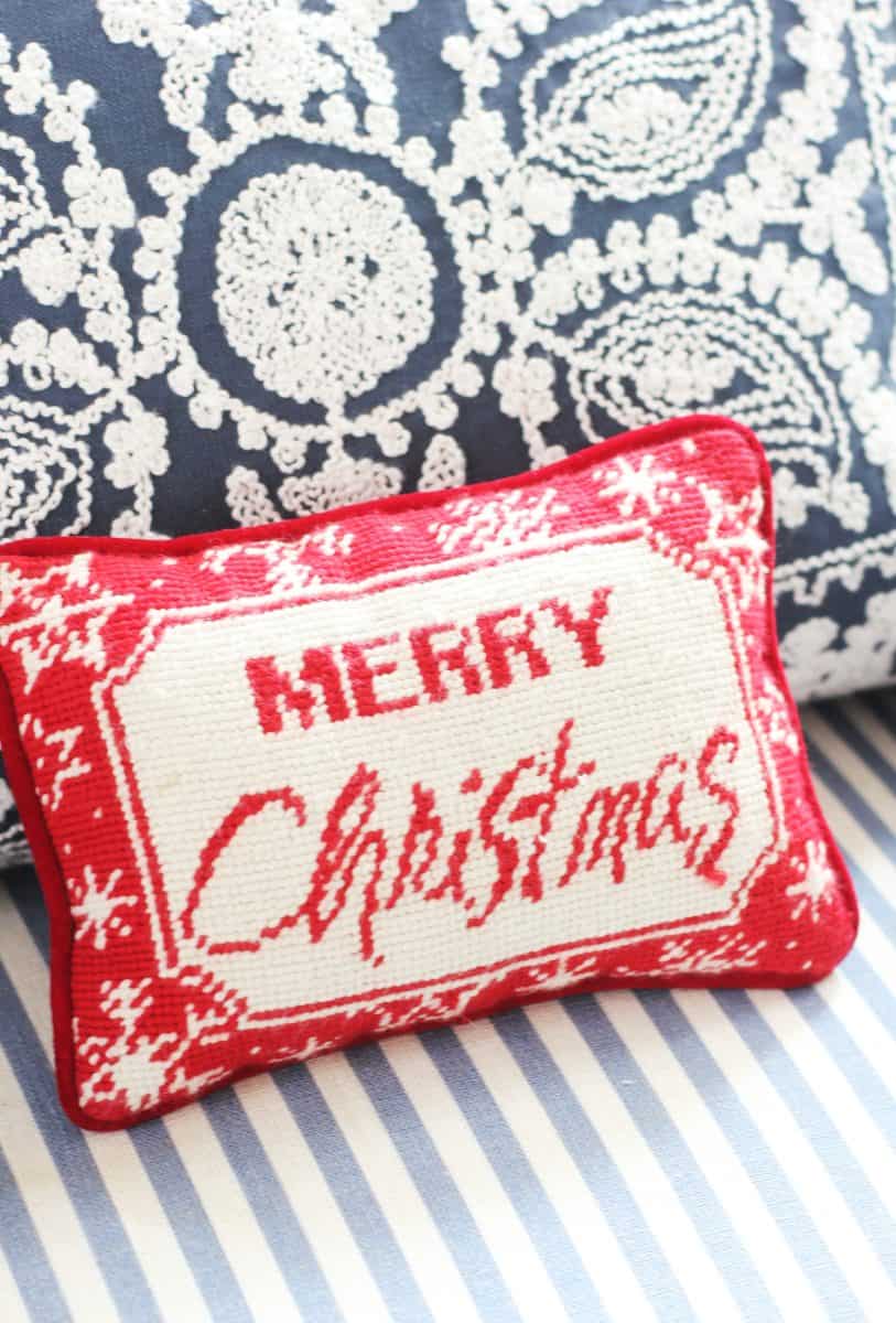 a small red and white decorative pillow with the words "merry christmas" stitched on it rests against a patterned blue and white cushion on a striped surface.