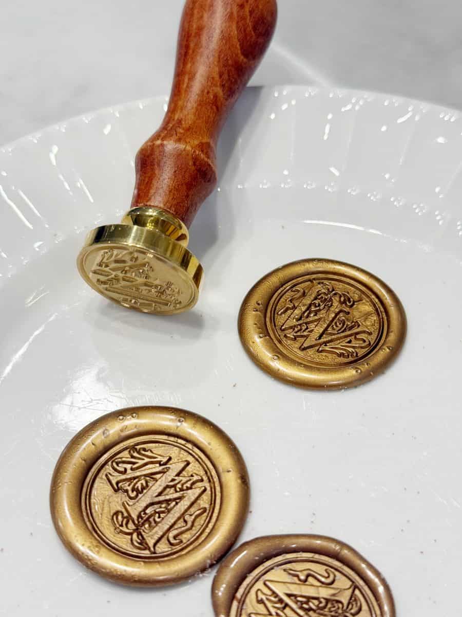 a wooden-handled wax seal stamp rests on a white plate next to three round, gold wax seals featuring an ornate letter "a" design.