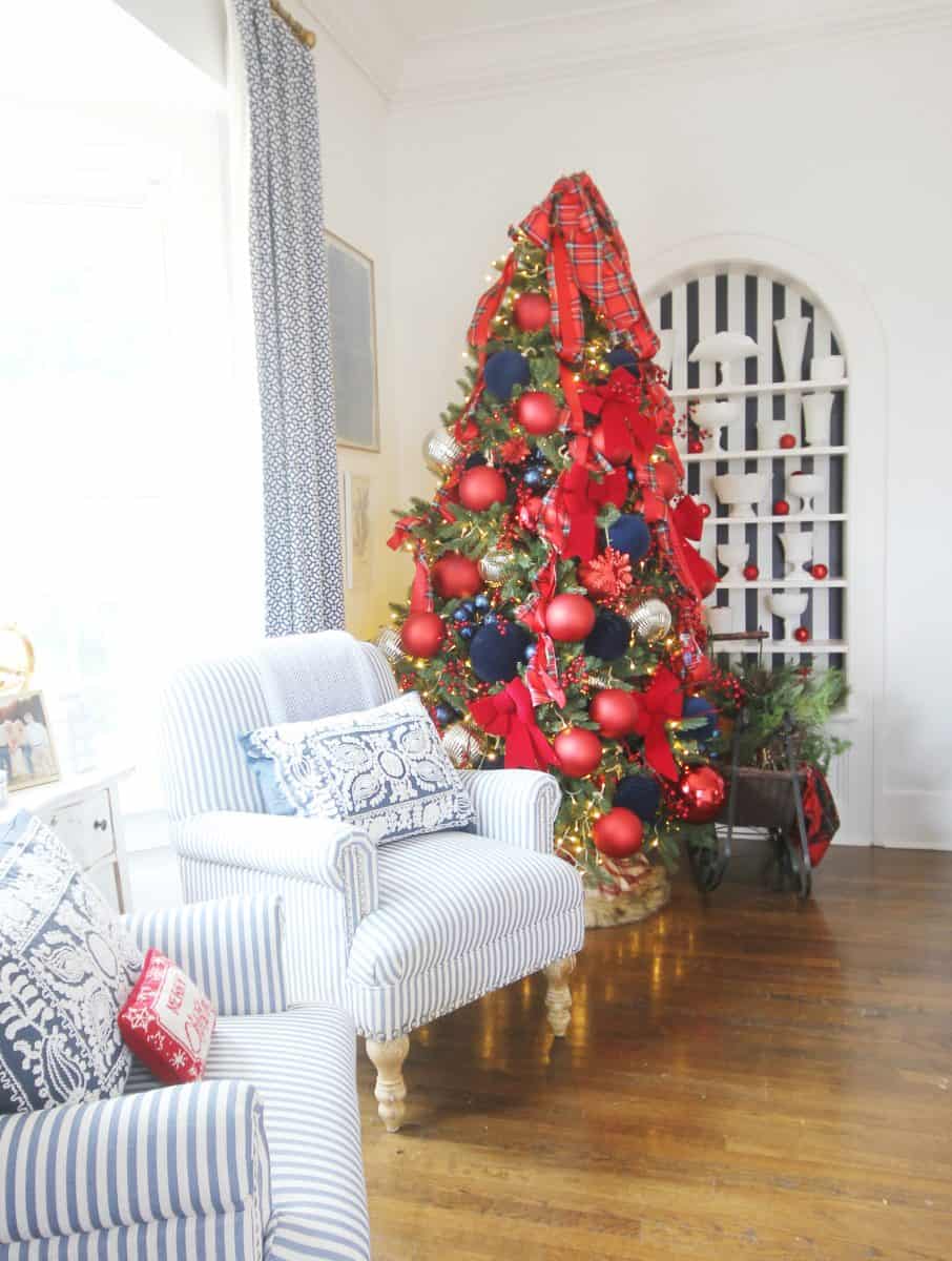 a christmas tree decorated with red bows, navy blue and red ornaments, and plaid ribbon stands in a bright living room with striped armchairs and a hardwood floor.