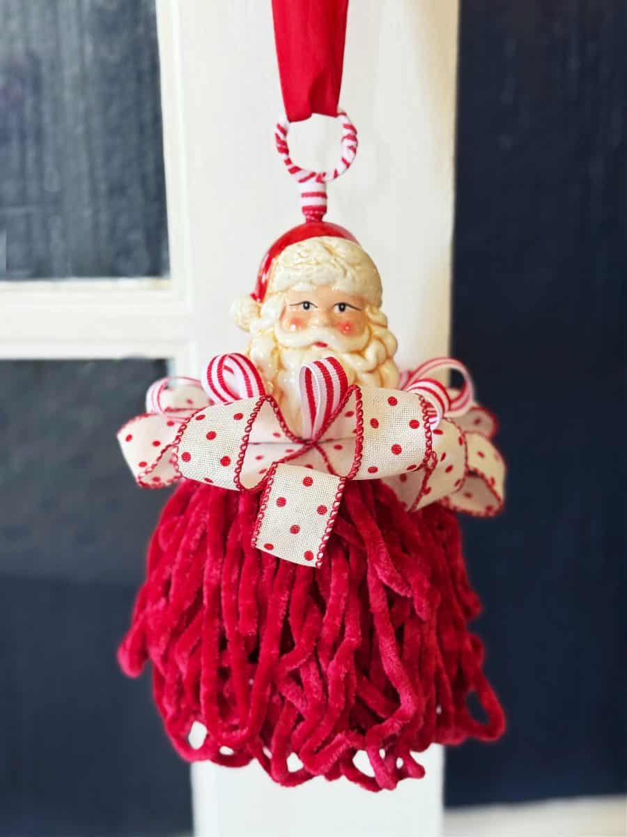 diy christmas tassel simple stepbystep tutorial 33 a santa claus ornament with a ceramic face, red and white ribbon bow like those made when learning how to make a giant bow, and a skirt of thick red yarn strands, hangs by a red ribbon in front of a window.