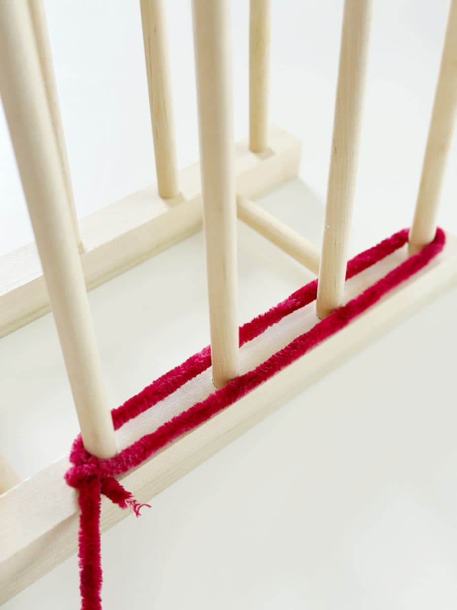 diy christmas tassel simple stepbystep tutorial 38 a close-up of a red pipe cleaner threaded through the wooden rods of a light-colored rack, resting on a white surface—an essential step in learning how to make a giant bow.