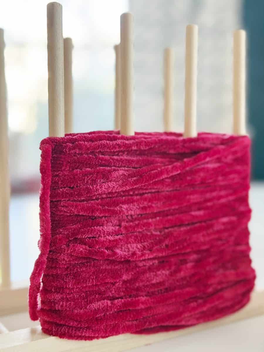 diy christmas tassel simple stepbystep tutorial 39 a close-up of several wooden dowels on a rack with plush, red velvet yarn wrapped around them, creating layers of soft texture—perfect inspiration if you're searching for how to make a giant bow. the background is softly blurred.