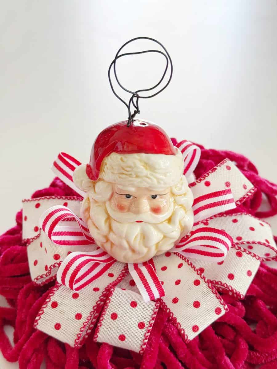 diy christmas tassel simple stepbystep tutorial 44 a santa claus ornament with a red hat and white beard sits on a large red and white polka dot ribbon bow, atop a textured red mat. perfect for those learning how to make a giant bow, the ornament has a wire loop for hanging.