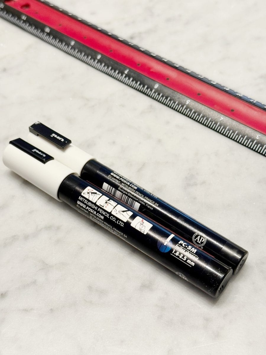 how to paint a gingerbread gift bag 27 two black marker pens with white caps are lying on a white marble surface next to a red and silver ruler. the markers are labeled with japanese text and brand information.