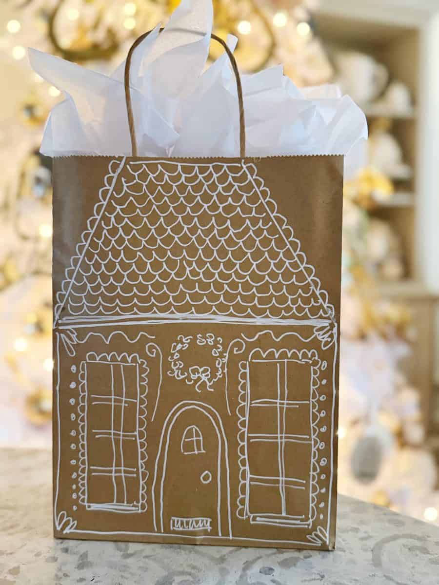 how to paint a gingerbread gift bag 26 a brown gift bag decorated with white drawings to resemble a gingerbread house, filled with white tissue paper. a blurred christmas tree with lights is visible in the background.