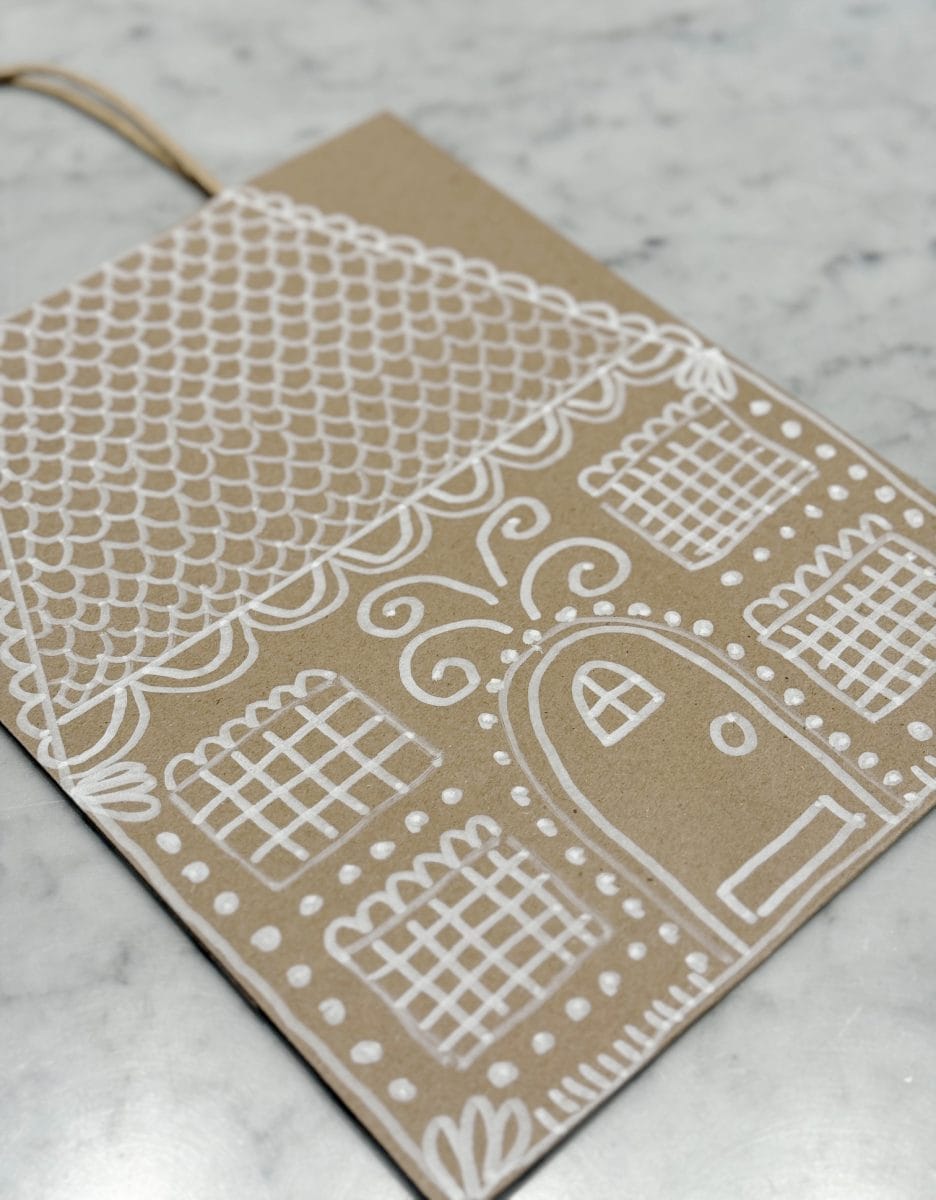 how to paint a gingerbread gift bag 23 a brown card decorated with a white, hand-drawn gingerbread house design, featuring a scalloped roof, grid windows, and a rounded door, lying on a light-colored marble surface.