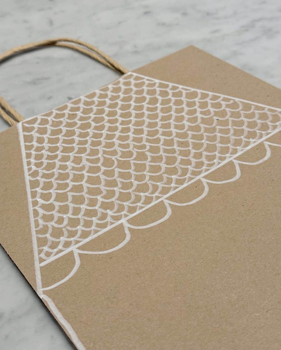 how to paint a gingerbread gift bag 30 a brown paper bag with twisted handles sits on a marble surface, featuring a hand-drawn white scallop pattern across the top half.