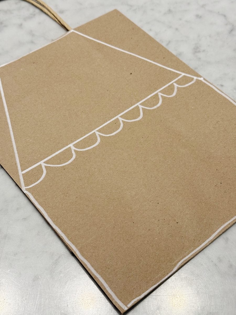 how to paint a gingerbread gift bag 28 a brown paper bag with white lines and scallop shapes drawn to resemble a storefront awning, placed on a light marble surface.