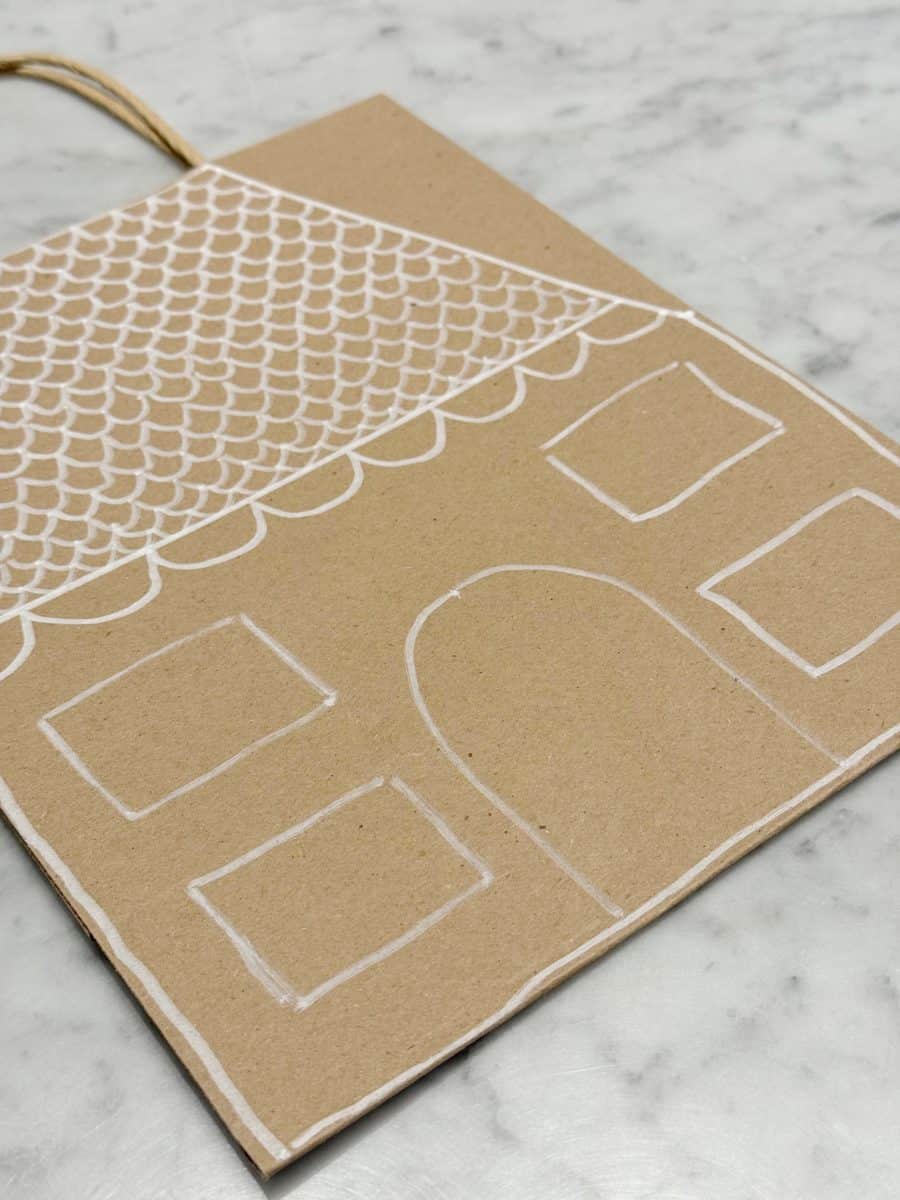 how to paint a gingerbread gift bag 31 a brown paper bag lies on a marble surface, decorated with a simple white line drawing of a house with a scalloped roof, four windows, and an arched door.