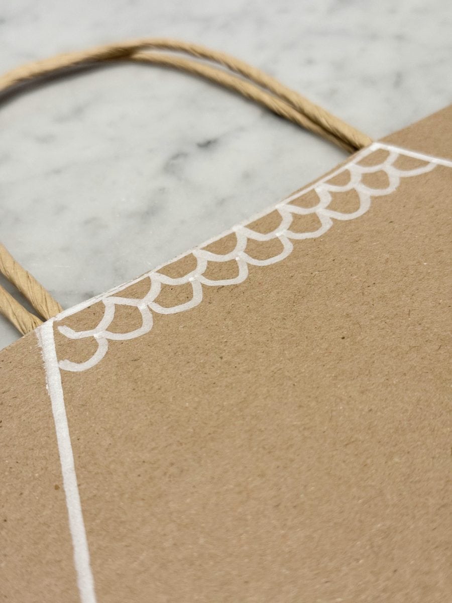 how to paint a gingerbread gift bag 29 a close-up of a brown paper bag with twisted paper handles, featuring a white hand-drawn scallop pattern along the edge, placed on a light gray marble surface.