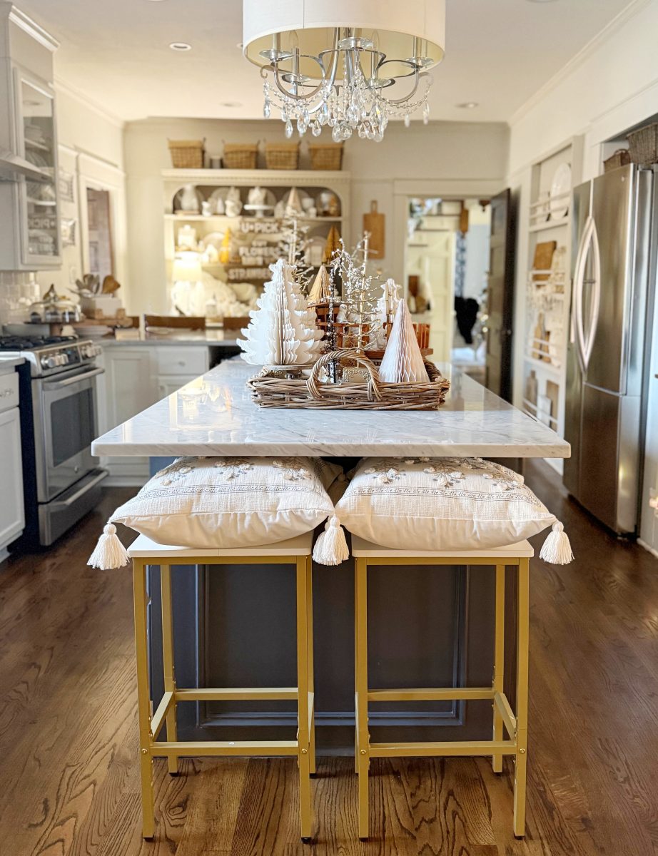 a cozy kitchen with a marble island, two cushioned stools, winter or holiday-themed decor on the counter, open shelves with dishes, and stainless steel appliances. warm wood floors and soft lighting create an inviting atmosphere.