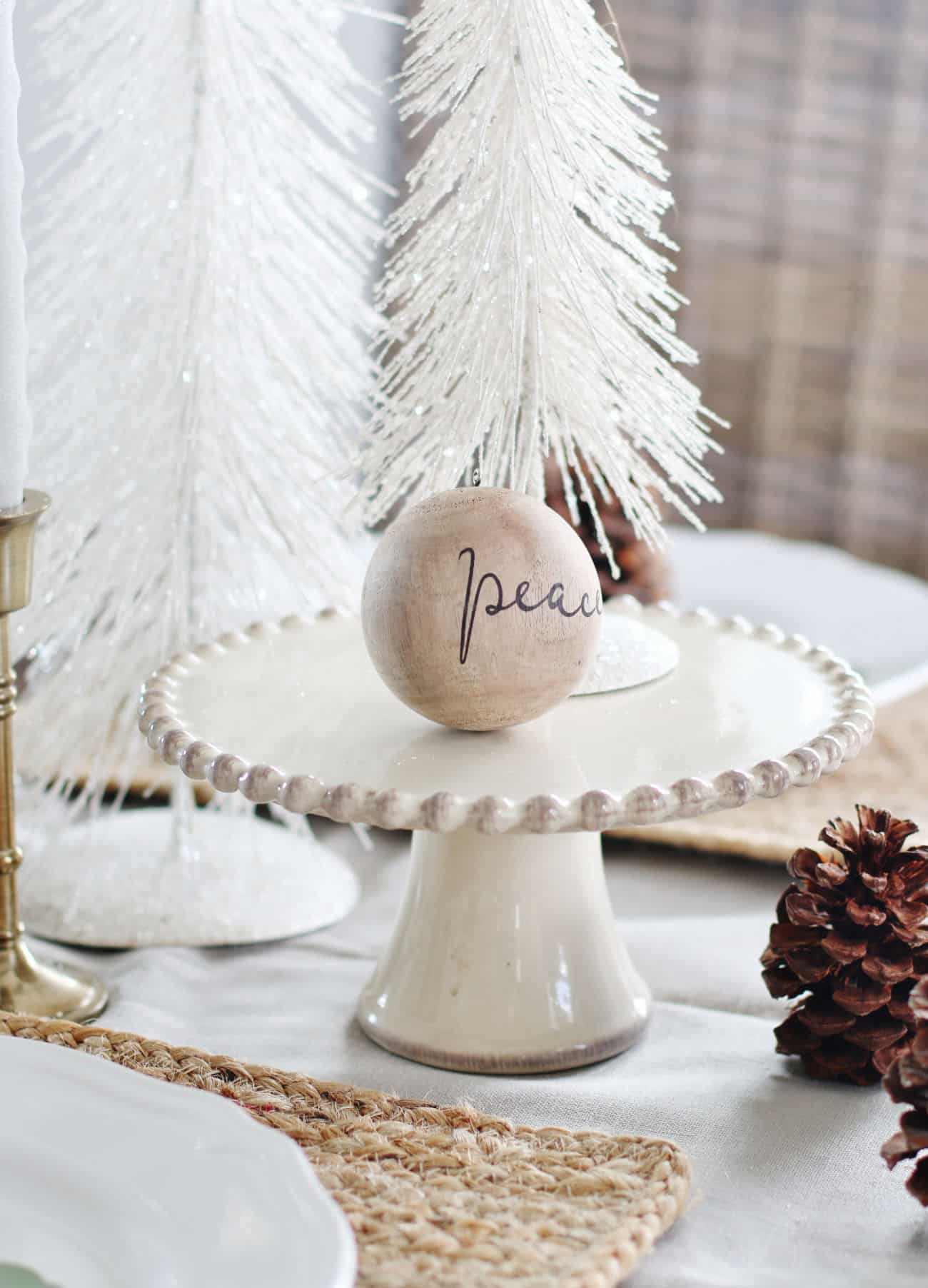the christmas ornament that started it all 22 a rustic holiday table setting featuring a wooden ornament with "peace" written on it, placed on a small white cake stand, surrounded by a white artificial tree and pine cones.