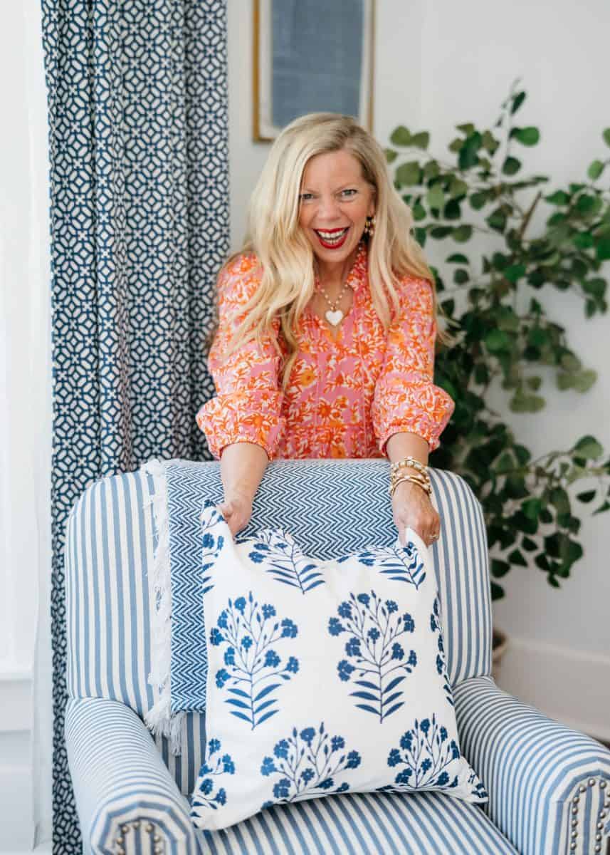 a smiling woman with long blonde hair and a bright floral dress places a white pillow with blue floral patterns on a striped blue armchair, with a leafy plant and patterned curtain in the background.