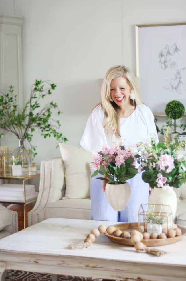a woman with long blonde hair, wearing a white blouse and light blue pants, smiles as she arranges pink flowers in a vase on a white coffee table in a bright, cozy living room decorated with plants.