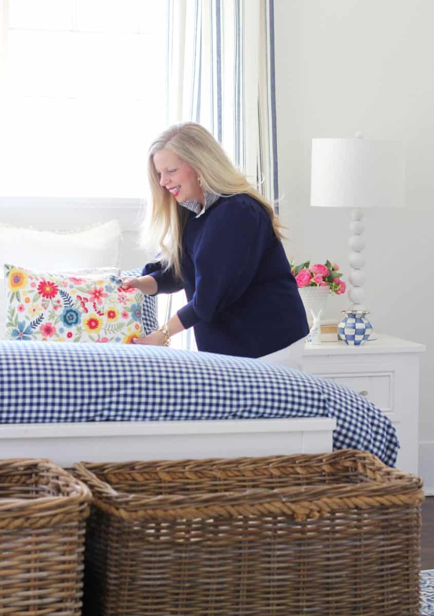 a woman with long blonde hair arranges pillows on a bed in a bright bedroom. the bed has a blue and white gingham comforter and colorful floral pillows. a white lamp and pink flowers are on the bedside table. there are wicker baskets at the foot of the bed.