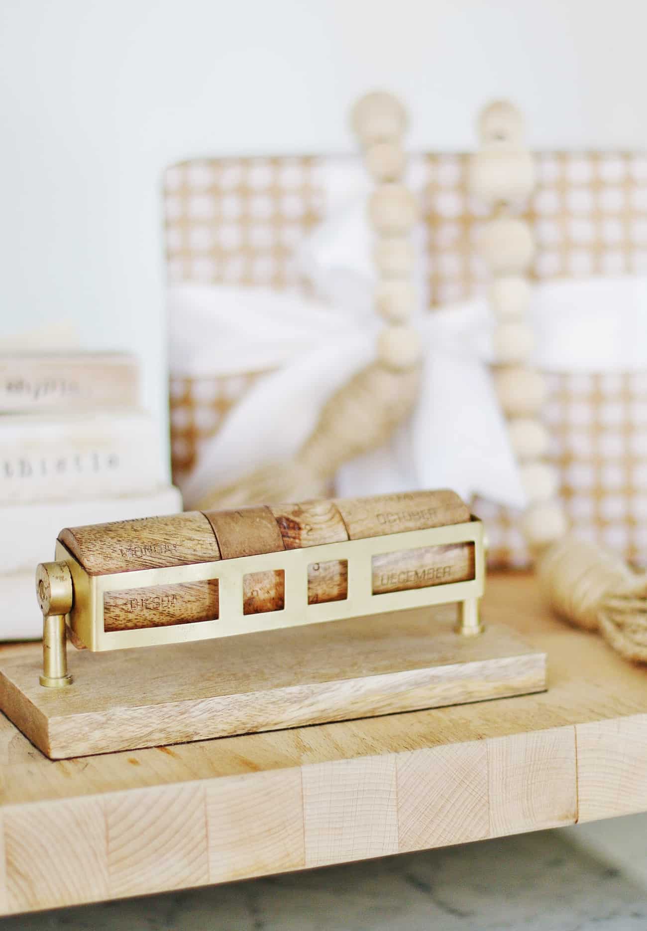 a wooden perpetual calendar with brass details on a desk, set against a backdrop featuring decorative books and geometric-patterned boards.
