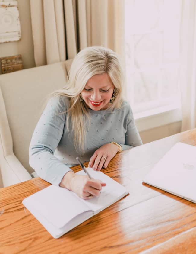 a woman with blond hair, smiling and writing in a notebook at a wooden desk, with a laptop beside her and curtains in the background.