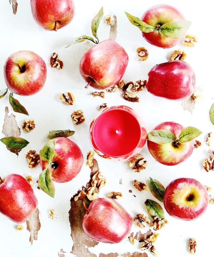 flat lay of fresh red apples, a red candle, walnuts, and leaves scattered against a white background.
