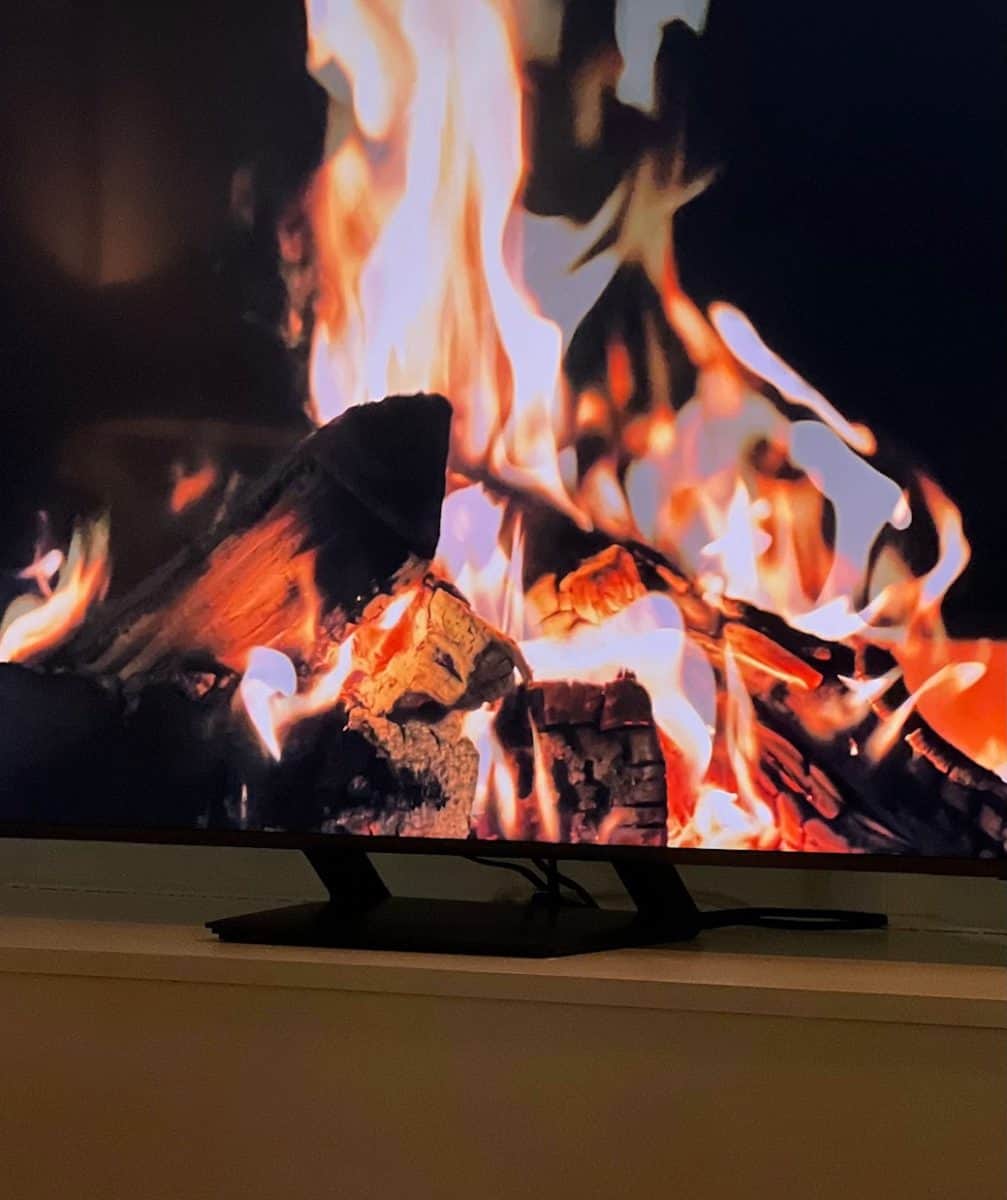a cozy, crackling fire burns brightly on a tv screen, displaying flames and embers in warm hues of orange and red. the tv is placed on a light-colored stand, creating a relaxing and inviting atmosphere.