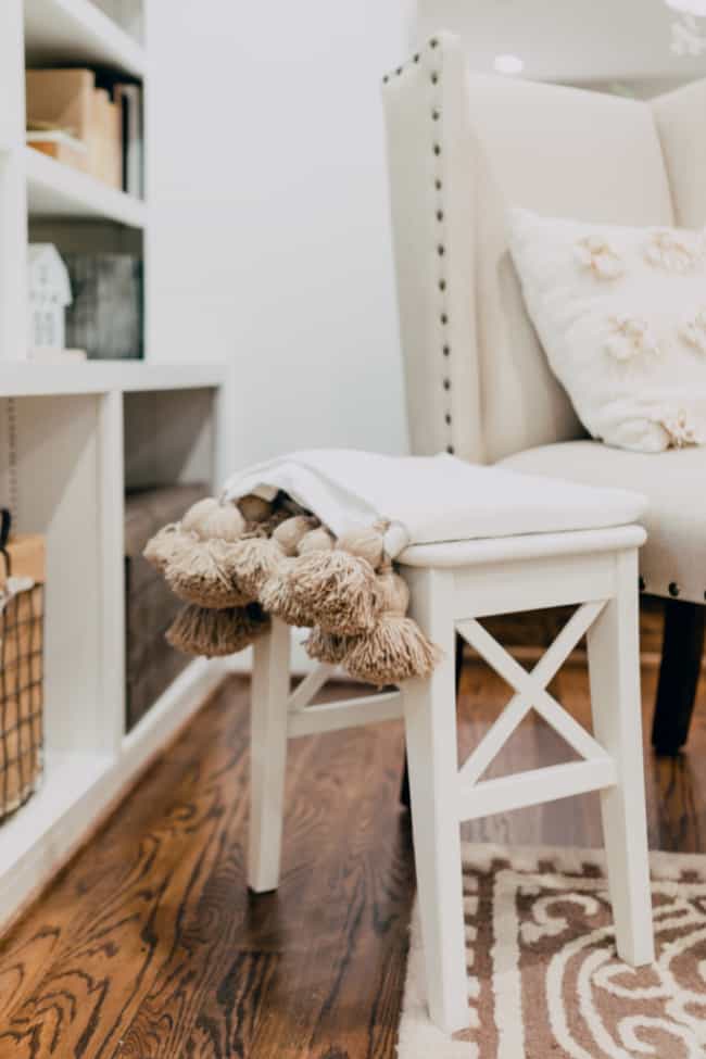 a cozy reading nook featuring a white ottoman with tassels on a throw blanket, adjacent to a white chair by a bookshelf, set on a hardwood floor with a patterned rug.
