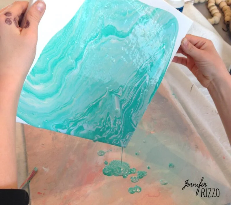 how to do acrylic paint pouring-for beginners 4 blue swirled paint being poured off of a canvas held by two. hands