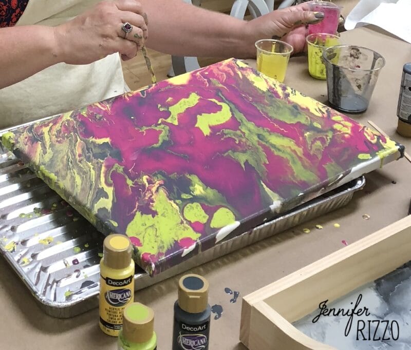 how to do acrylic paint pouring-for beginners 5 a hand swirling brightly paint after paint pouring with a stick. the paint is bright colors and is surrounded by cups of paint and paint bottles